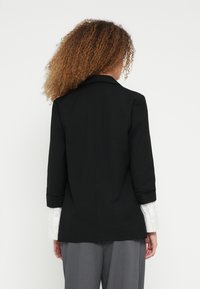 Μαύρο σακάκι με δομημένο σχέδιο, με μακριά μανίκια, μία πίσω σχισμή και λευκό πουκάμισο που διακρίνεται κάτω από τα μανίκια.