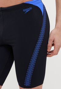 Shorts de bain noirs avec des accents bleus, présentant un motif en zigzag sur le côté et un logo Speedo sur la taille. Fabriqués en tissu lisse et élastique.