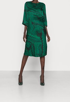 Femme portant une robe verte mi-longue avec des motifs noirs tourbillonnants et des talons verts assortis, debout les jambes écartées sur un fond blanc.
