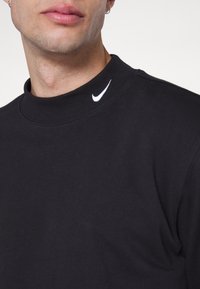 Černé tričko s dlouhým rukávem a vysokým límcem, s malým bílým logem Nike swoosh na levé straně. Materiál vypadá hladký a měkký.