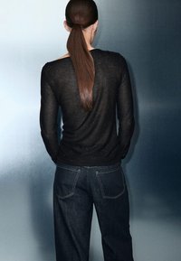 Black knitted sweater with a textured finish, paired with dark blue high-waisted jeans. Model's hair is tied back, facing away.