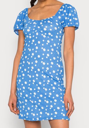 Robe bleue avec motif de marguerites blanches, manches courtes à volants, décolleté carré et coupe ajustée. Le tissu texturé offre des détails de côtes subtils.