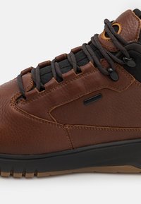 Botte de randonnée en cuir marron avec lacets noirs, col rembourré et semelle texturée noire et beige, vue en gros plan de côté.