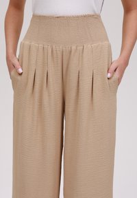 Beżowe spodnie szerokie z teksturowanym wykończeniem, z marszczonym wysokim stanem i bocznymi kieszeniami. Materiał wydaje się lekki i swobodny.