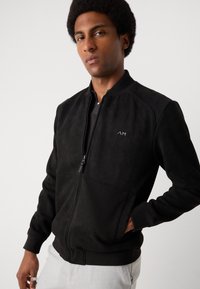 Black suede bomber jacket with a front zipper, ribbed cuffs, and a small logo on the chest. Features a smooth texture and fitted design.