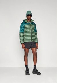 Polstret grønn og blågrønn jakke med hette, glidelåslukking og The North Face-logo. Brukes med svarte shorts og en matchende lue.