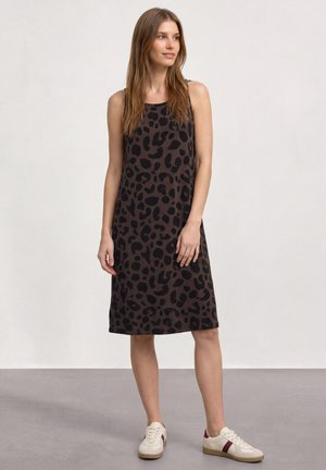 Femme portant une robe marron foncé sans manches avec des taches léopard noires, debout dans des baskets blanches avec des rayures bordeaux sur un sol gris.