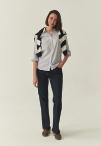 Chemise à rayures avec col et boutons, blanche et bleue, associée à un jean foncé. Pull rayé noir et blanc posé sur les épaules, chaussures marron.