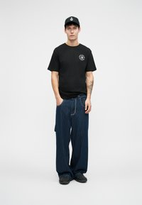 Zwarte katoenen t-shirt met een ronde graphic op de borst, gecombineerd met blauwe gestreepte wijde broek en donkere schoenen. Een zwarte pet maakt de look compleet.