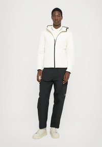 Hvitt quiltet jakke med hette, svart glidelås, sammen med svarte bukser med sidelommer og logo. Modellen bruker beige sneakers.