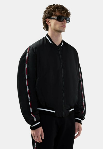 Mężczyzna z kręconymi włosami, ubrany w czarną kurtkę na zamek z biało-czerwonym paskiem z logo GCDS na rękawach oraz pasujące czarne spodnie, okulary przeciwsłoneczne, na białym tle.