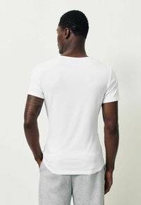 Fehér pamutpóló, kerek nyakkivágással és rövid ujjakkal, testhezálló szabás, hátulról látható, szürke melegítőnadrággal párosítva.