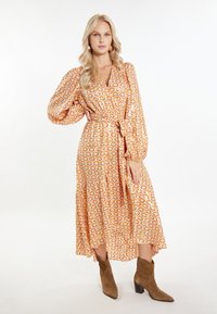Rochie în stil wrap din țesătură portocalie, cu modele florale, mâneci lungi tip puff, curea în talie și lungime midi. Este asortată cu botine maro.