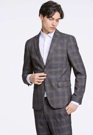 Bruin geruit pak met blauwe accenten, enkele knoopsluiting, met notched revers en twee zijzakken. Draagt over een wit shirt.