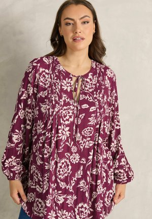 Kobieta w fioletowej, kwiecistej bluzce z długim rękawem, z dekoltem w kształcie otworu i wiązaniem, stojąca na tle jednolitego jasnego tła.