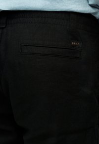 Κοντινό πλάνο της πίσω τσέπης μαύρου παντελονιού με ραμμένη γλώσσα και μικρό ορθογώνιο λογότυπο BOSS πάνω από αυτήν.