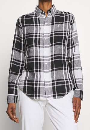 Personne portant une chemise à carreaux noire, blanche et grise à boutons avec manches longues et un pantalon blanc sur un fond uni.