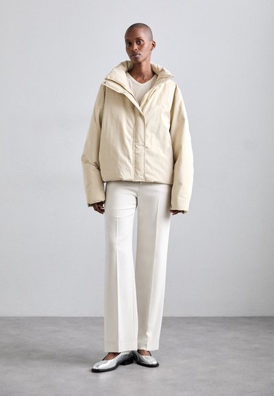 Kreemjas puffjakis kõrge krae, vabalt lenduv lõige ja täis esikuclosure, koos valgete pükste ja hõbedaste kingadega tekstuurse põranda peal.