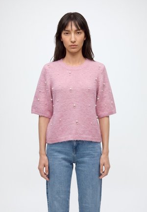 Femme portant un pull à manches courtes rose clair avec de petites perles blanches et un jean bleu, debout devant un fond uni.