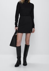 Un pull col roulé noir associé à une mini-jupe noire avec des fentes sur les côtés, des bottes en cuir jusqu'aux genoux et un sac fourre-tout noir.