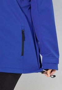 Blå mjukskaljacka med en dragkedjeficka, justerbar nederkant med snörstoppar och smooth yta. Har en figurnära design och långa ärmar.