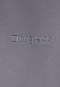 Grijs katoenen t-shirt met zwarte geborduurde tekst "Dropsize" in een decoratief lettertype met scherpe hoeken en elegante rondingen.