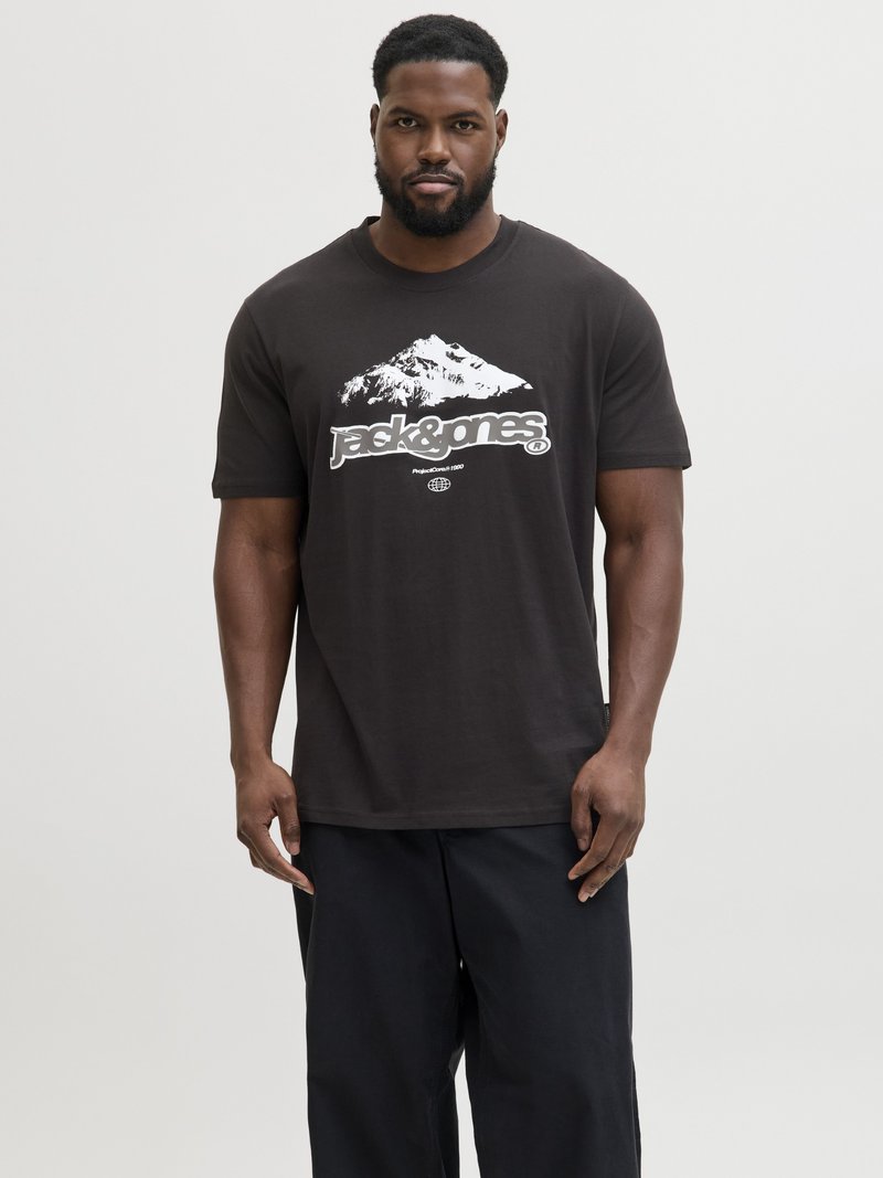 Man draagt zwart T-shirt met witte bergafbeelding en tekst "jack&jones", gecombineerd met losse zwarte broek, staand tegen een effen achtergrond.