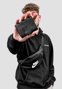 Black waist bag with a smooth texture and Nike logo, worn over a black hoodie. The person is holding a black wallet in their hand.