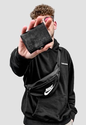 Black waist bag with a smooth texture and Nike logo, worn over a black hoodie. The person is holding a black wallet in their hand.