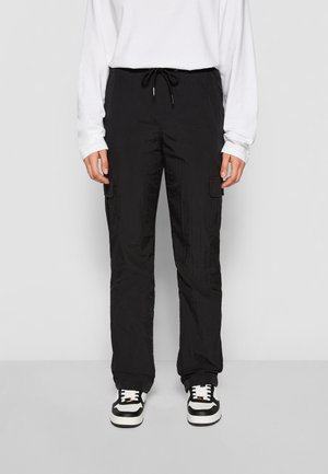 Personne portant un pantalon cargo noir avec taille à cordon, chemise blanche à manches longues et baskets noir et blanc, debout devant un fond uni.