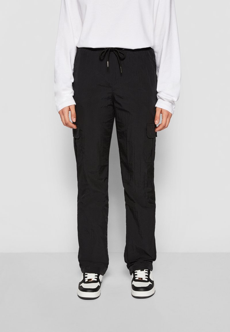 Personne portant un pantalon cargo noir avec taille à cordon, chemise blanche à manches longues et baskets noir et blanc, debout devant un fond uni.