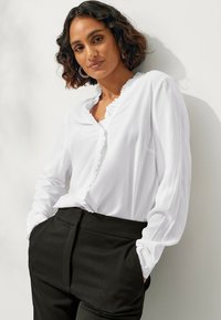 Witte blouse met ruches aan de halslijn en voorkant, lange mouwen en knoopmanchetten, gecombineerd met hoogopgetrokken zwarte broek. Glad stoftextuur.