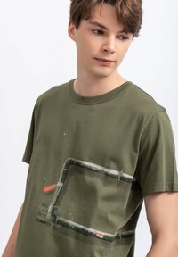 T-shirt en coton vert olive avec un graphique imprimé d'un tuyau rehaussé de touches rouges et de texte. Manches courtes, col rond, coupe décontractée.