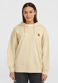 Beżowa bluza z kapturem o luźnym kroju, z ribbowymi mankietami i okrągłą naszywką z logo na piersi. Miękka tekstura i minimalistyczny design.