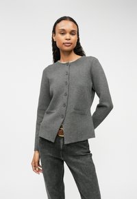 Gilet en tricot gris avec un col arrondi, sept boutons à l'avant et deux poches avant. Il a une coupe ajustée et une texture lisse.