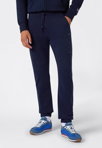Marineblauwe trainingsbroek met elastische boorden en een trekkoord in de taille; heeft subtiele logo-details langs de zijkant; gecombineerd met blauwe en grijze sneakers.