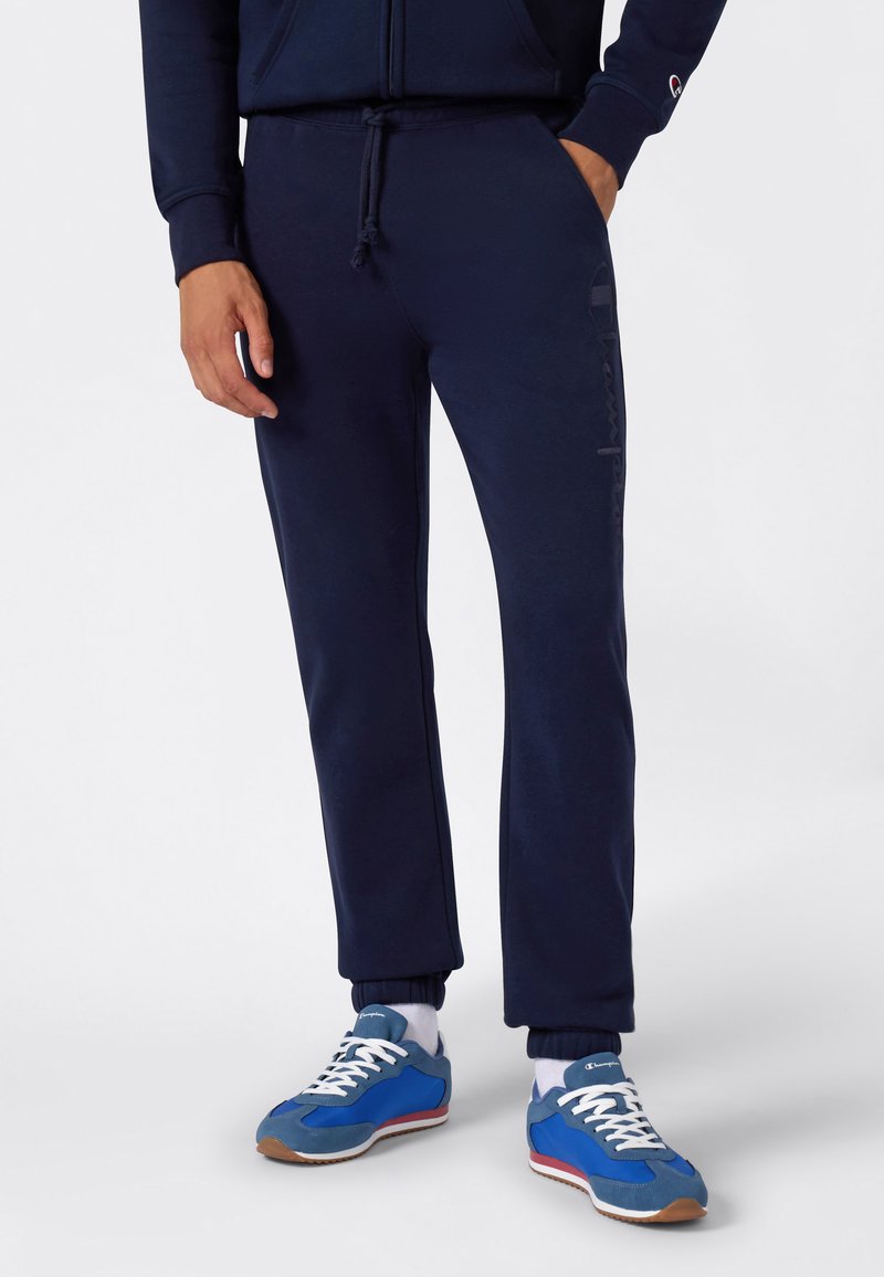 Marineblauwe trainingsbroek met elastische boorden en een trekkoord in de taille; heeft subtiele logo-details langs de zijkant; gecombineerd met blauwe en grijze sneakers.