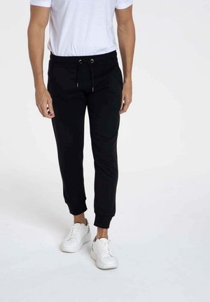 Pantalons de jogging noirs en tissu doux, dotés d'une taille élastique avec cordon de serrage, de poignets côtelés et de poches latérales. Portés avec des baskets blanches.
