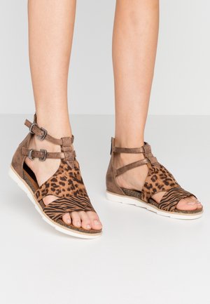 Voeten in open sandaaltjes met bruine dierenprint riemen, dubbele enkelgespen en witte zolen op een effen lichte achtergrond.