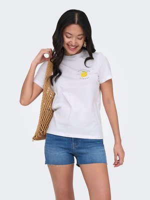 Mujer sonriente con camiseta blanca con diseño de limón, shorts vaqueros, sosteniendo un bolso de hombro tejido color camel, de pie contra un fondo liso.