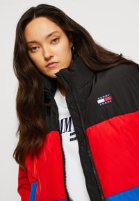 Červená, čierna a modrá puffová bunda s vysokým golierom, zipovým zapínaním a logom Tommy Jeans. Vyrobená z lesklého, izolačného materiálu.