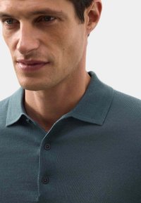 Polo en bleu sarcelle, avec un tricot texturé, deux boutons au col et un design de col classique. Gros plan sur fond neutre.