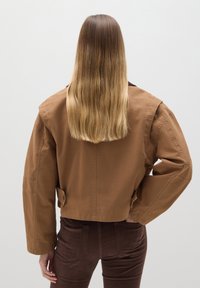 Braune cropped Jacke mit weiten Ärmeln, einem Knopfdetail auf der Rückseite, kombiniert mit dunkelbraunen Cordhosen. Glatte Textur, minimalistisches Design.
