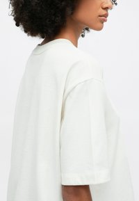 Valge ülisuur T-särk, mis on valmistatud pehmest puuvillakangast. Omab ümarat kaelust ja lühikesi varrukasid, mille otstes on keeratud ääred.