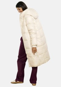 Krēmskrāsas pūkains mētelis ar kapuci, ar quiltējošu dizainu. Brīvs piegriezums, garums sasniedz līdz celim, apvienots ar bordo biksēm.