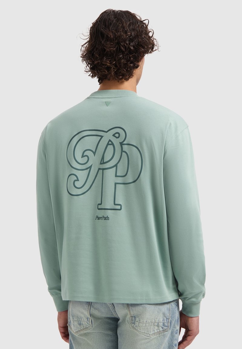 Persoon met krullend haar, gekleed in een groene langemouwen shirt met grote "PP" en "PurePath" op de achterkant, in combinatie met lichte spijkerbroek.
