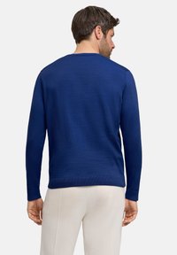 Marineblauer Pullover mit geripptem Halsausschnitt und Bündchen, aus strukturiertem Stoff gefertigt, getragen mit hellfarbenen Hosen. Die Rückansicht zeigt eine enganliegende Silhouette.