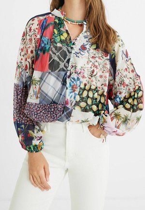 Kobieta w wielokolorowej, kwiecistej bluzce typu patchwork z długimi bufkami, w połączeniu z białymi spodniami z wysokim stanem i warstwową biżuterią z koralików.