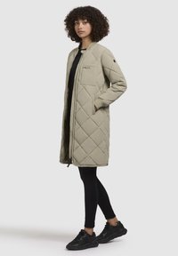 Cappotto imbottito trapuntato di un beige chiaro con zip frontale, tasche laterali e un design semplice. Abbinato a leggings neri e sneakers.