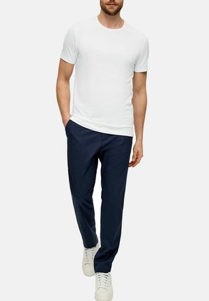 Mann trägt ein einfaches weißes kurzärmliges T-Shirt, marineblaue Hose und weiße Sneakers, steht mit einer Hand in der Tasche.